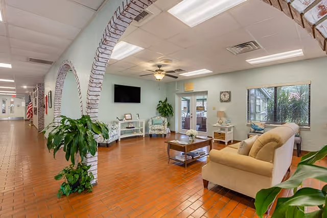 Bright common lounge area with sofas, armchairs, coffee table, wall-mounted TV, arched brick columns, and potted plants.