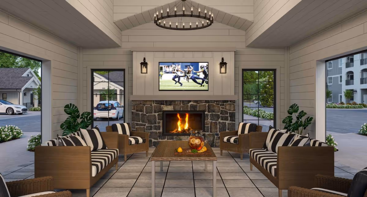 A cozy indoor lounge area with a stone fireplace and a flat-screen TV mounted above it showing a football game. The room features wooden furniture with black and white striped cushions arranged around a rectangular coffee table with a pitcher of iced tea and glasses. Large windows on three sides provide views of parked cars and surrounding buildings. A circular chandelier hangs from the ceiling.