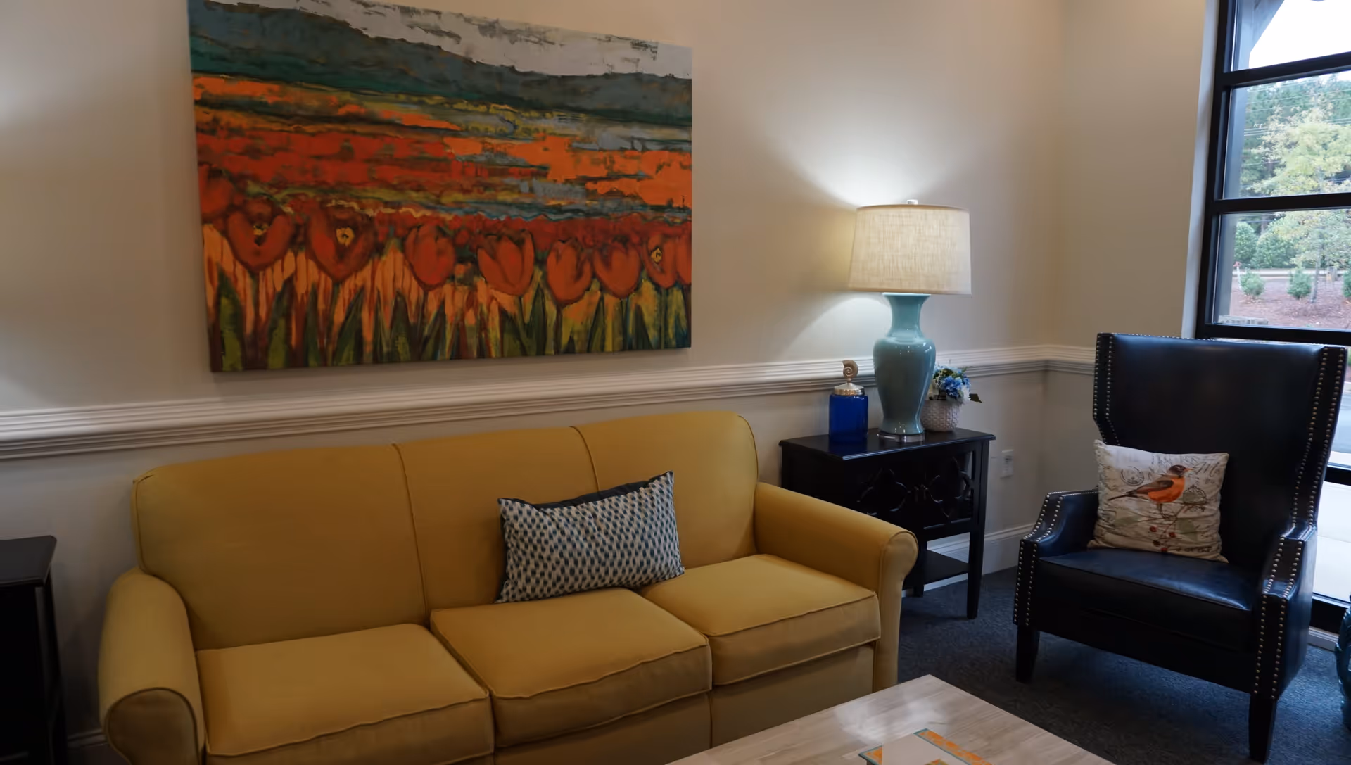 A cozy living room area featuring a yellow sofa with a patterned cushion, a black armchair with a bird-themed pillow, a side table with a blue lamp and decorative items, a large colorful painting on the wall, and a window showing greenery outside.