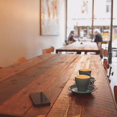A long wooden dining table with cups on saucers and a smartphone, in a bright dining area with a few people seated near the window in the background.
