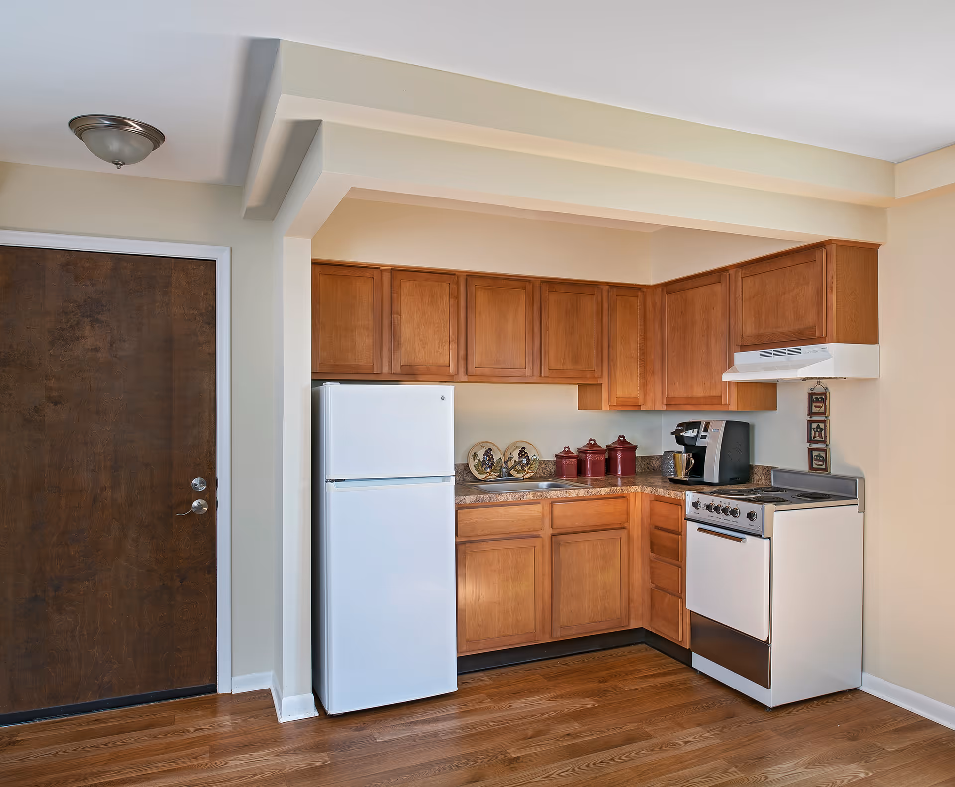 A small kitchen area with wooden cabinets, a white refrigerator, a white stove with an oven, a coffee maker, and decorative items on the countertop. The floor is wooden, and there is a dark brown door to the left.