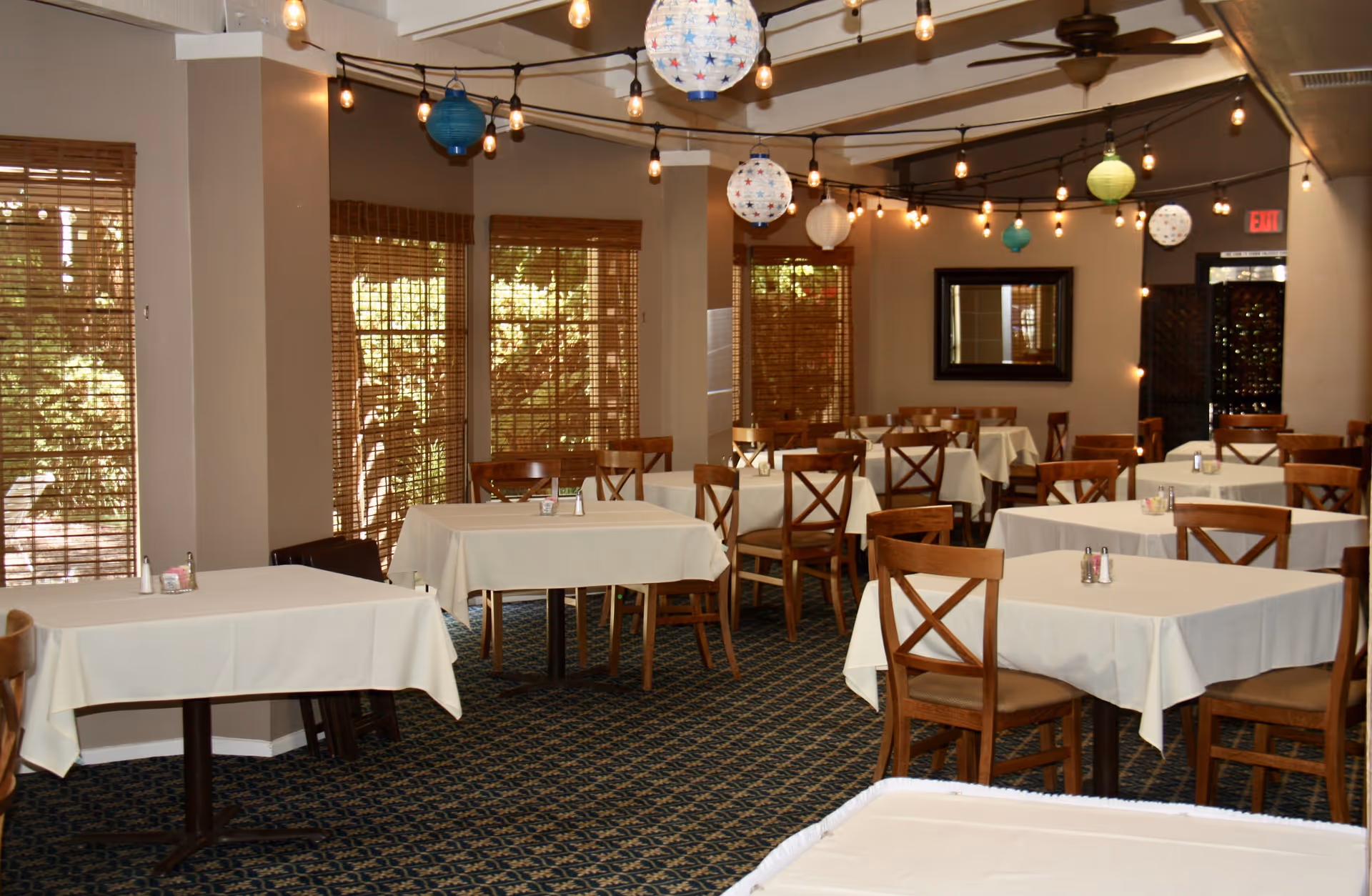 Dining room with multiple tables covered in white tablecloths and wooden chairs. The room has windows with wooden blinds, string lights with colorful paper lanterns hanging from the ceiling, and a mirror on the far wall.