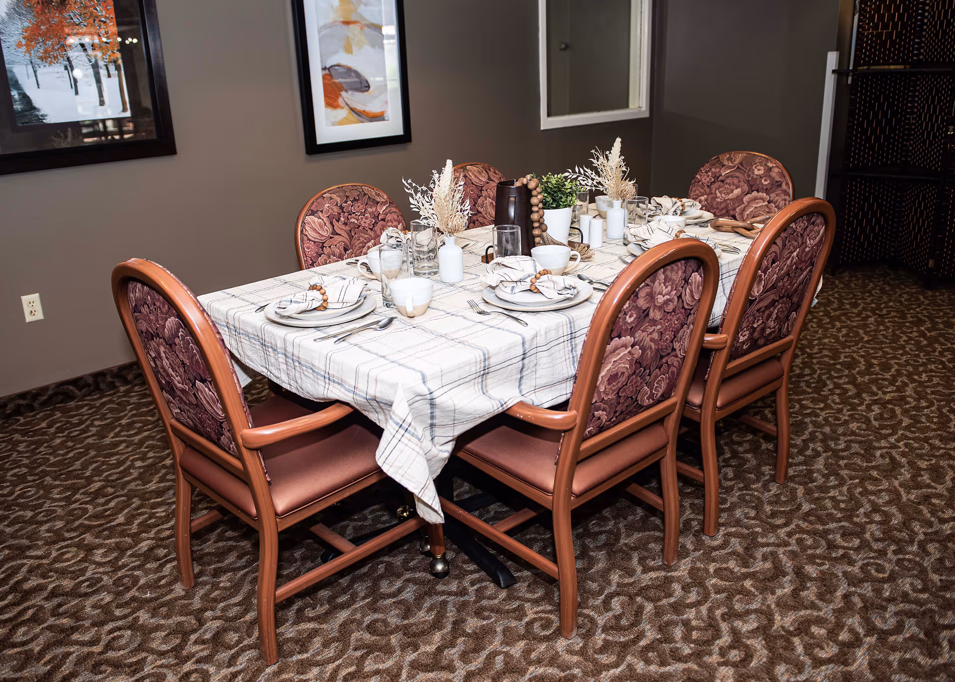 A dining table set for six with a plaid tablecloth, decorative place settings and floral-upholstered chairs in an interior dining room.