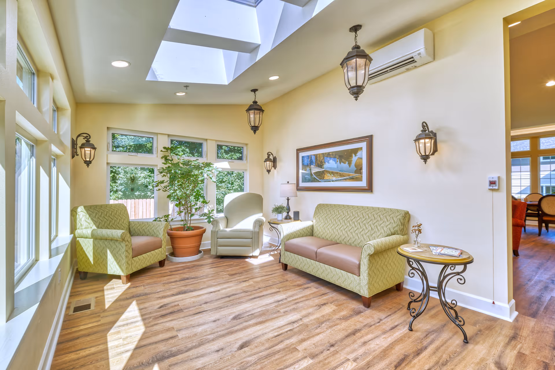 Sunlit seating area with green upholstered armchairs and a sofa, a potted plant, hanging lanterns, skylights, and wood flooring.