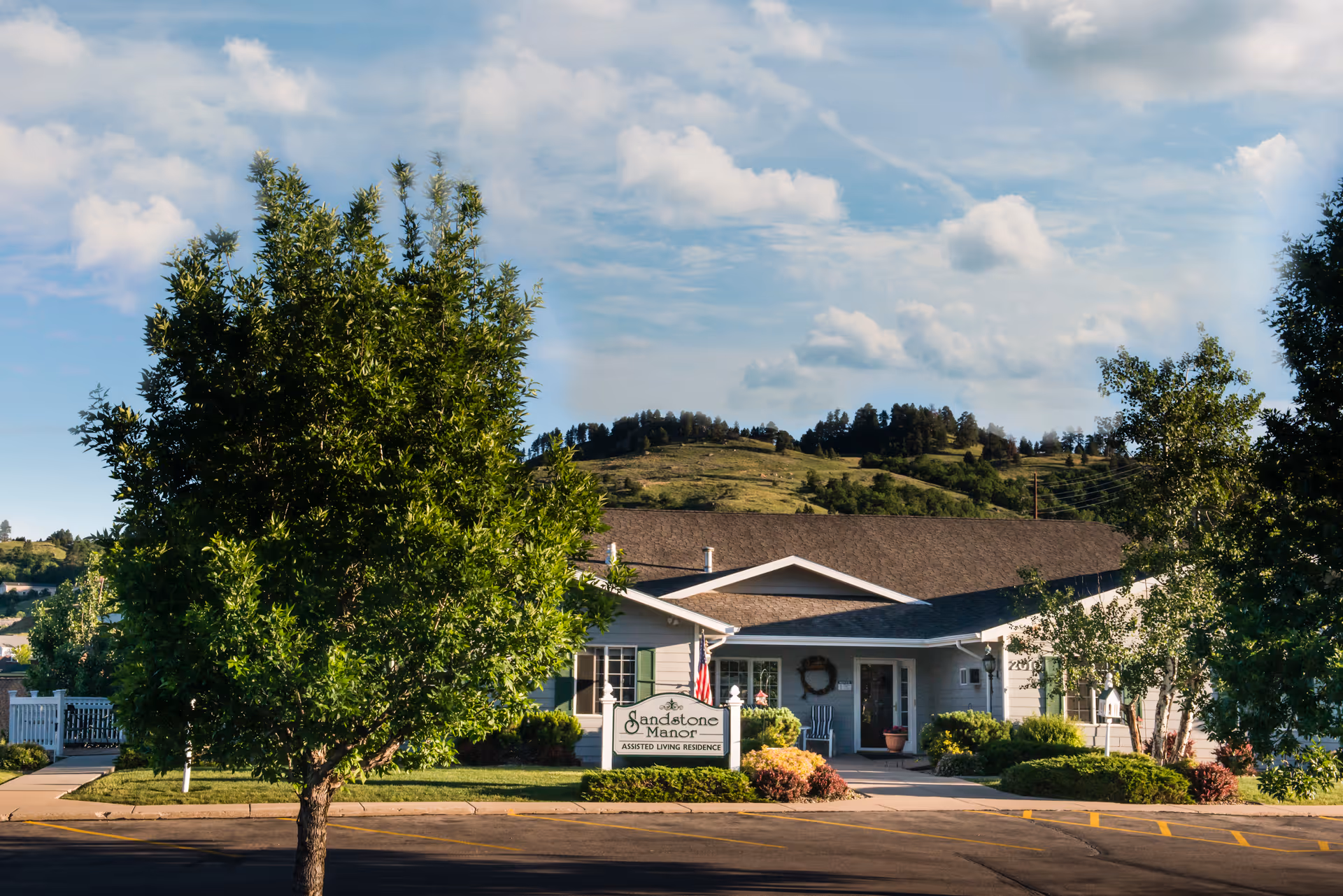 Front exterior of the Sandstone Senior Living building with a sign, trees, and a driveway under a partly cloudy sky.