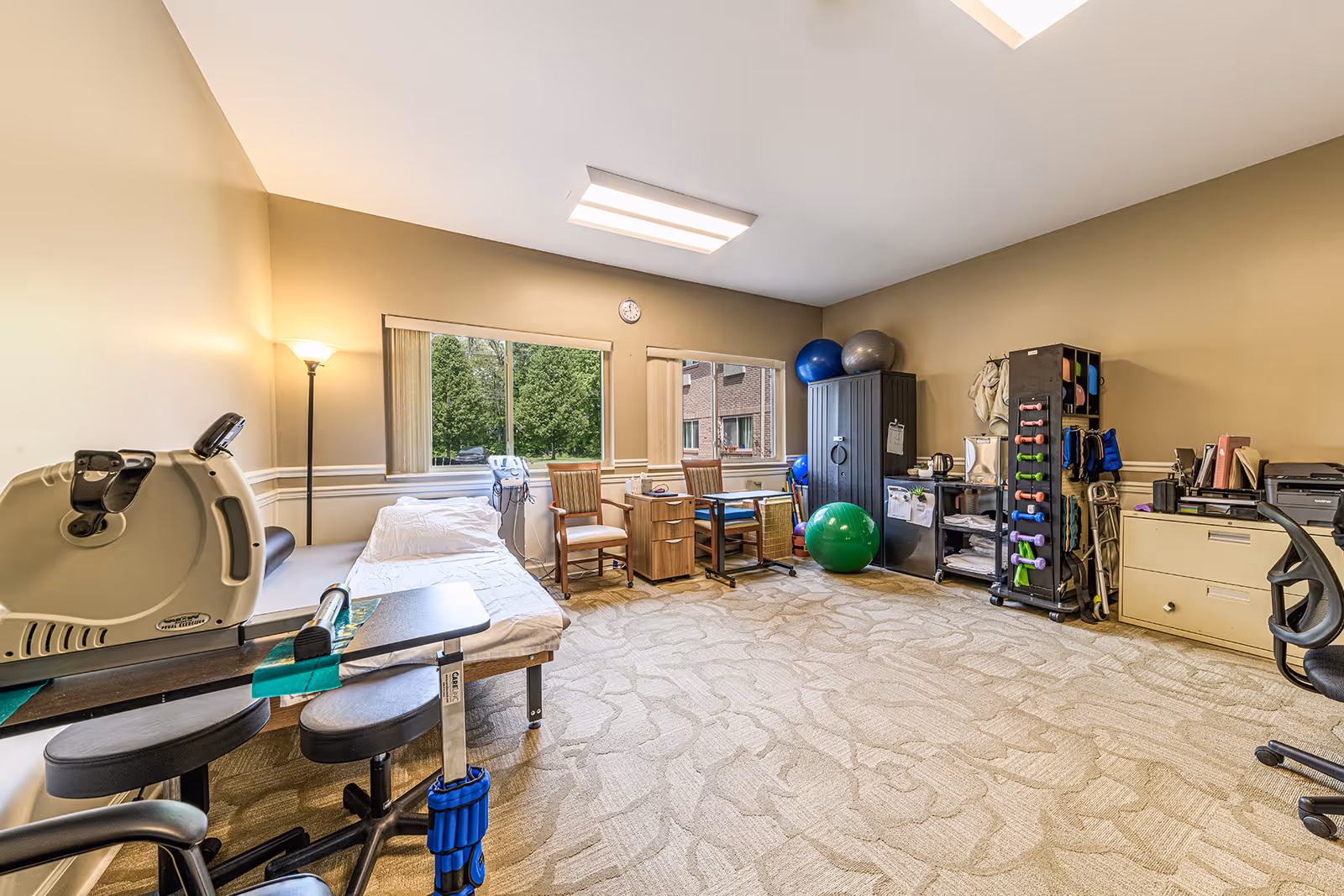 A well-lit therapy or rehabilitation room with exercise equipment including a pedal exerciser, therapy balls, dumbbells, and chairs. The room has beige walls, carpeted floor, two windows with vertical blinds, and various storage cabinets and desks.