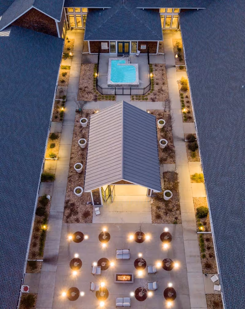Aerial view of an outdoor courtyard at dusk featuring a small building with a metal roof, a fenced hot tub, multiple potted plants, illuminated pathways, and a seating area with chairs arranged around a fire pit.
