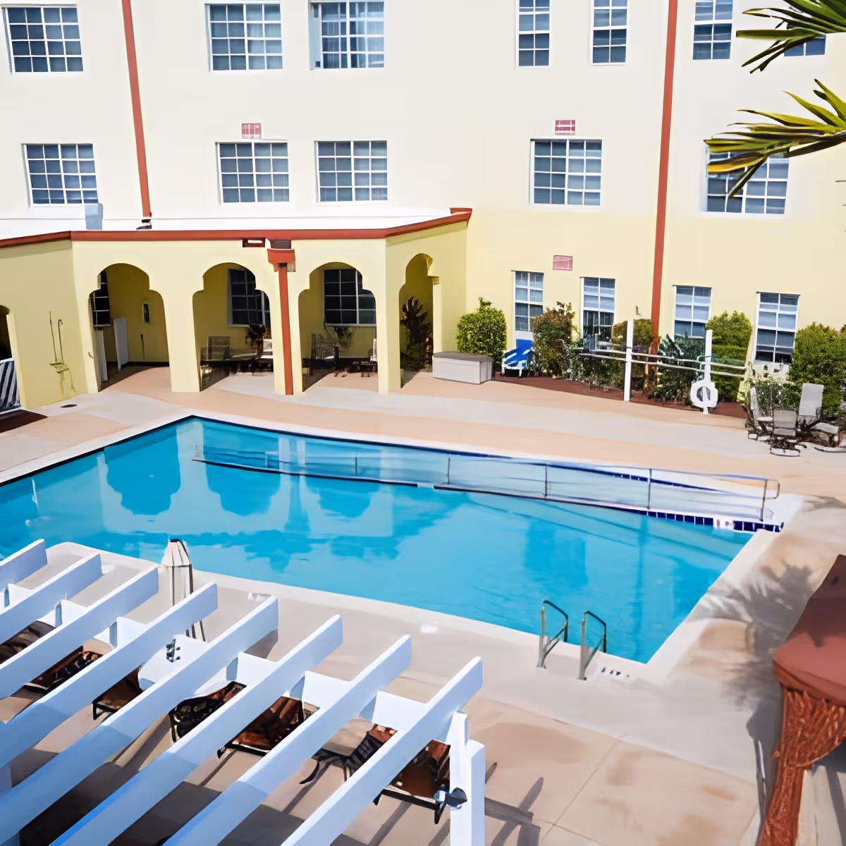 Outdoor swimming pool area at Discovery Village At Melbourne with clear blue water, surrounded by a beige deck. There are white pergolas with lounge chairs underneath, a yellow building with multiple windows in the background, and some greenery along the building walls.