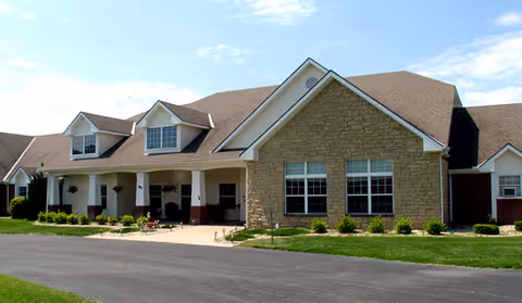 Exterior view of a single-story senior living facility building with a stone facade, multiple windows, and a covered entrance with white columns. The building is surrounded by a well-maintained lawn and a paved driveway under a partly cloudy sky.