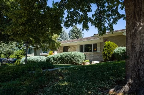 Single-story brick building front of an assisted living facility partially shaded by large trees with shrubs and a lawn.