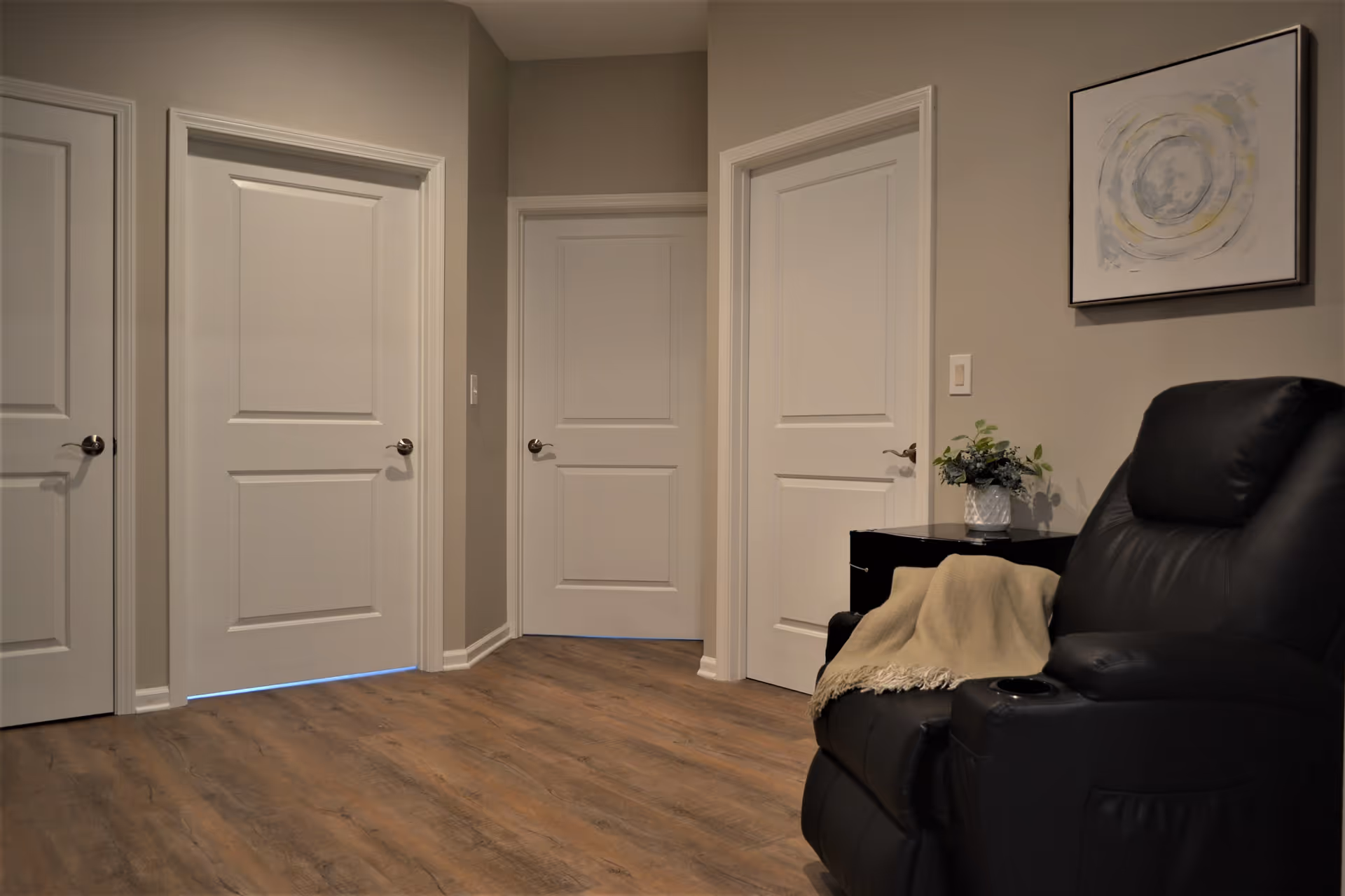 Interior view of a hallway with four closed white doors, light brown wooden flooring, beige walls, a black leather recliner chair with a beige throw blanket, a small black side table with a potted plant, and a framed abstract painting on the wall.
