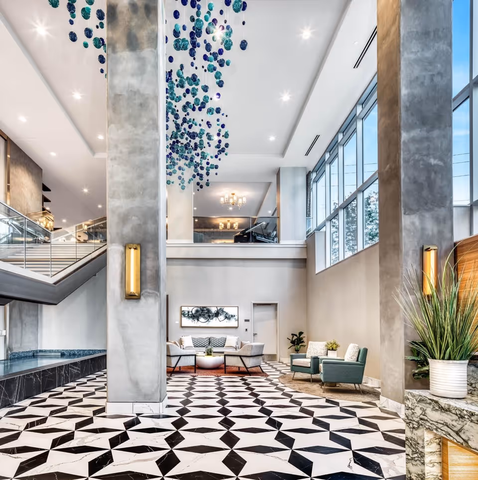 A spacious and modern senior living facility lobby with high ceilings, large windows, and geometric black and white tiled flooring. The area features comfortable seating with sofas and armchairs, decorative plants, and a unique hanging art installation made of blue and teal glass spheres. There is a staircase with glass railings on the left side and a grand piano visible on the upper level.