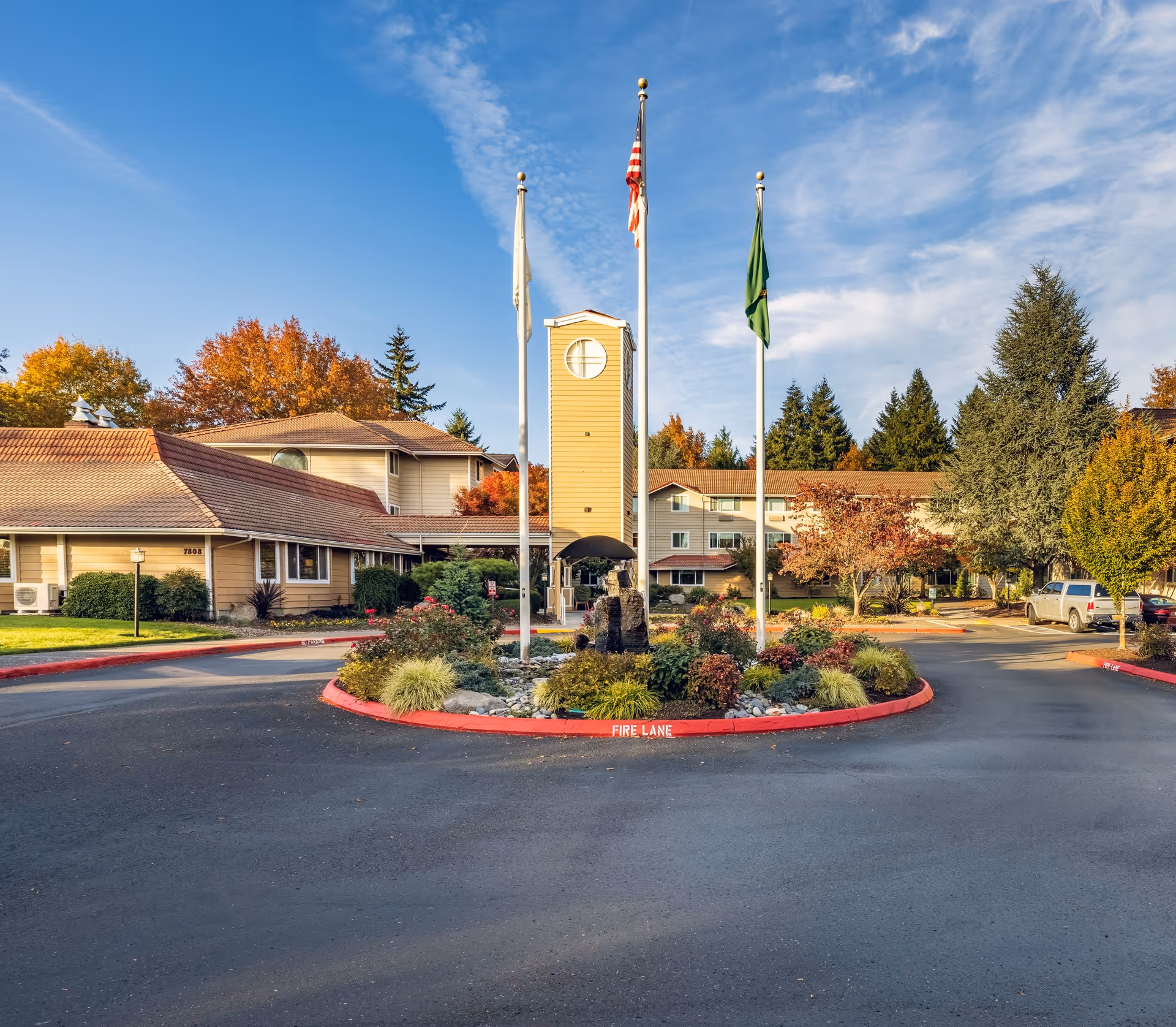 Exterior view of Van Mall Retirement Vancouver facility with a circular driveway, landscaped garden in the center with three flagpoles, and a clock tower structure. The building has beige siding and a brown roof, surrounded by trees with autumn foliage under a partly cloudy sky.