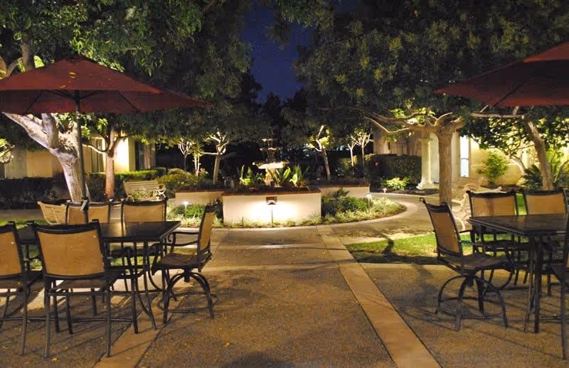 Outdoor patio area at night with metal tables and chairs under large red umbrellas, surrounded by trees and illuminated landscaping with a central fountain.