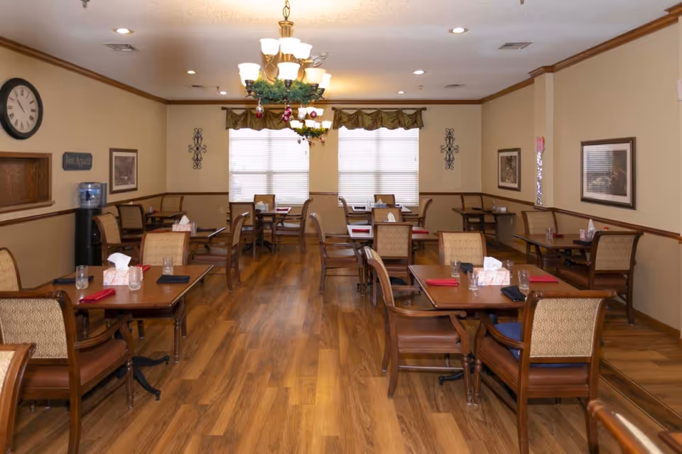 A dining room in a senior living facility with multiple wooden tables and chairs arranged neatly. Each table has glasses, napkins, and tissue boxes. The room has wooden flooring, beige walls with framed pictures, a clock, and a water dispenser. A chandelier decorated with greenery and ornaments hangs from the ceiling. Two large windows with blinds and valances allow natural light into the room.