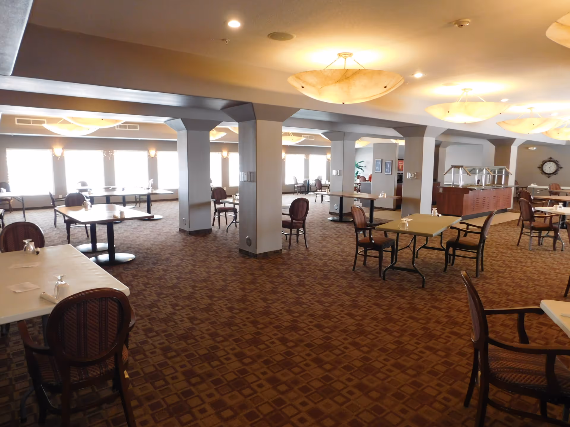 Spacious dining room with multiple tables and chairs arranged neatly. The room has large windows letting in natural light, carpeted floor with a patterned design, and ceiling lights providing warm illumination. There is a serving station along one wall and decorative elements such as a wall clock and framed pictures.
