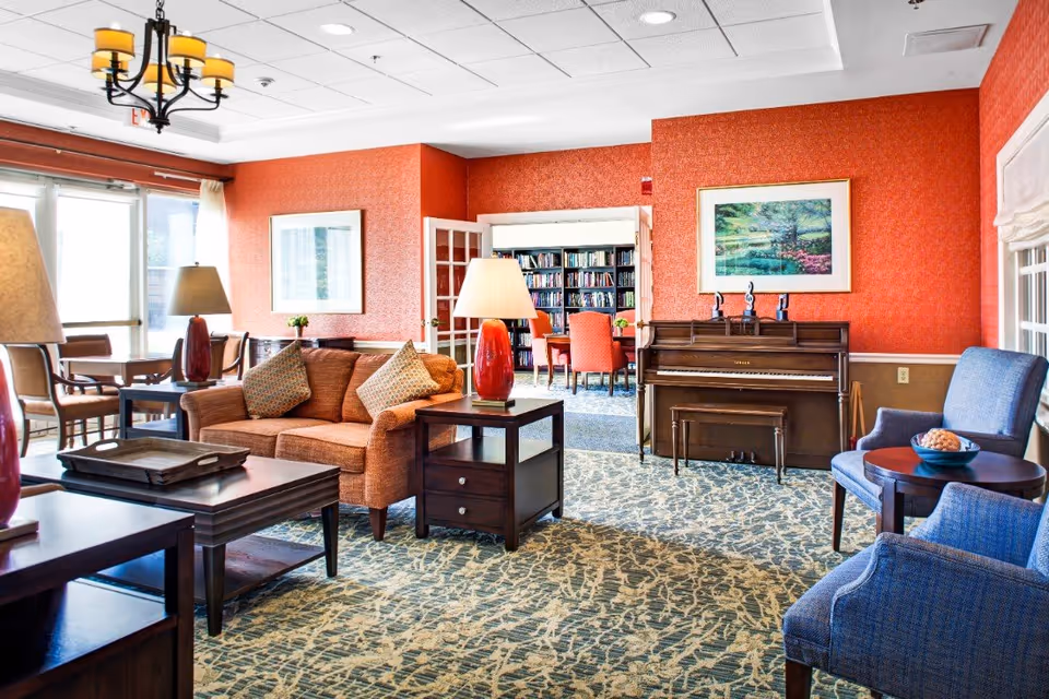 A cozy living room area in Bedford Court featuring an orange sofa with patterned cushions, two blue armchairs with a small round table, a wooden piano against a red textured wall, and a bookshelf visible through a doorway. The room is well-lit with natural light from large windows and has a patterned carpet and ceiling lights.