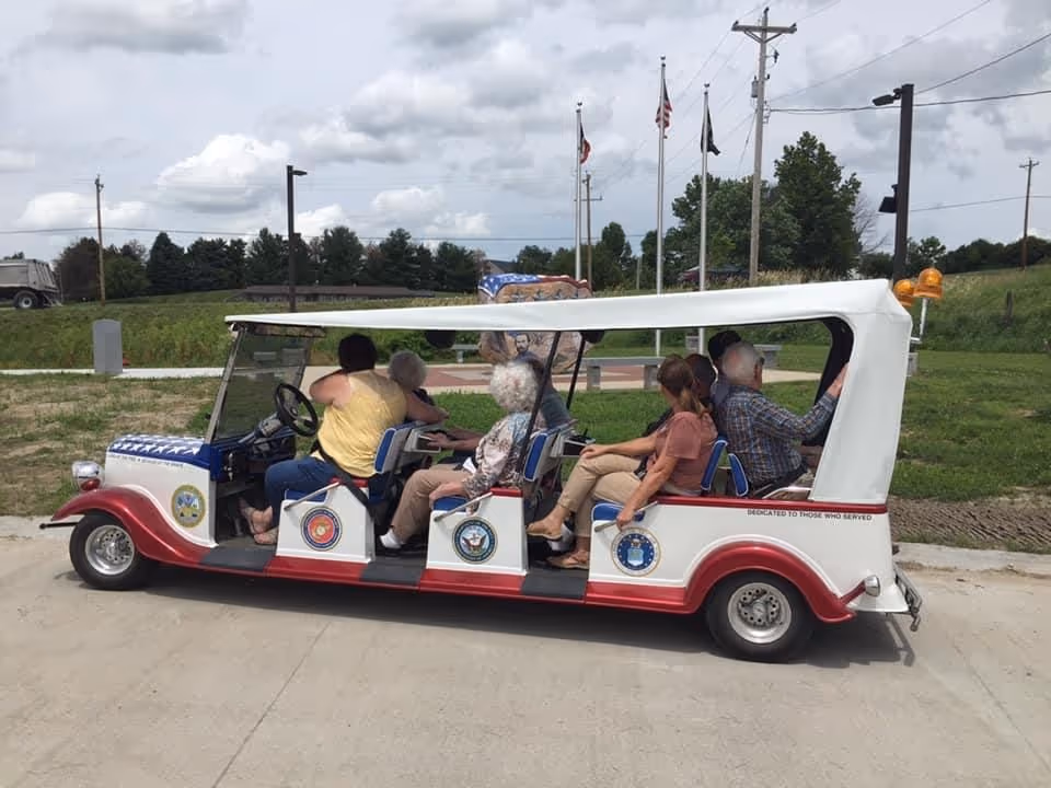 A group of elderly people riding in a white and red open-sided electric cart decorated with military service emblems, parked on a paved path with grass and flags in the background under a cloudy sky.