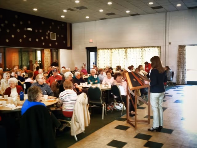 A large group of older adults seated at round tables in a community dining hall listening to a woman speaking at a podium.