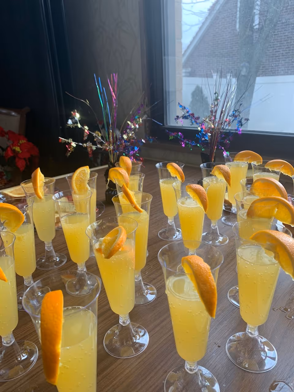 Multiple clear plastic glasses filled with a yellow beverage, each garnished with a slice of orange, arranged on a wooden table near a window with decorative colorful tinsel in the background.