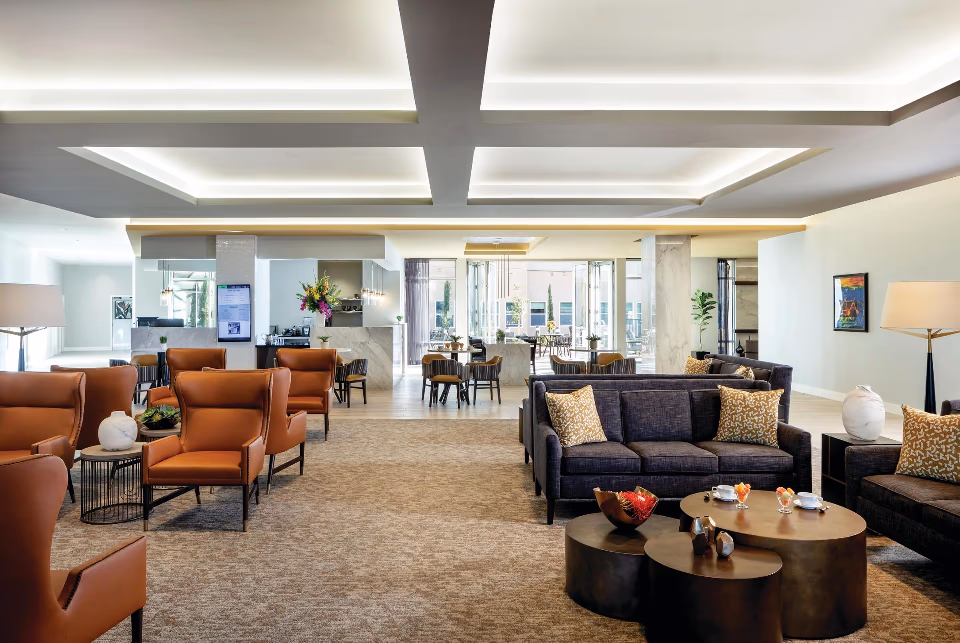 A spacious and modern living room area in a senior living facility featuring multiple brown leather armchairs and dark gray sofas with patterned cushions. The room has a beige carpet, round coffee tables with decorative items and cups, large windows letting in natural light, and a ceiling with recessed lighting. In the background, there is a dining area with tables and chairs and a counter with flowers and a coffee machine.