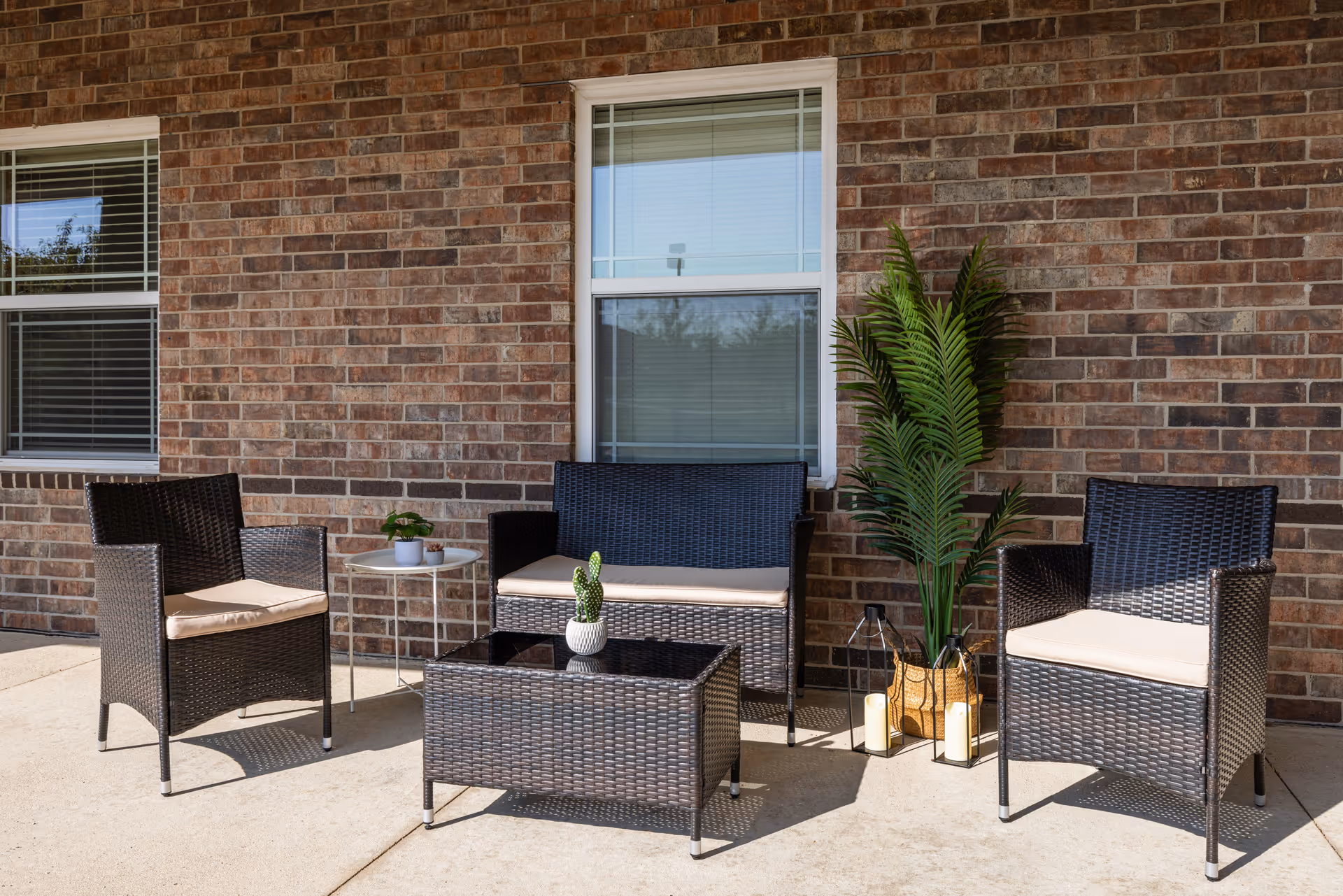 Outdoor patio area with a brick wall background featuring two black wicker chairs with beige cushions, a matching loveseat, a black wicker coffee table with a small potted cactus, a small round white side table with two small potted plants, and a tall green potted plant next to two lanterns with candles.