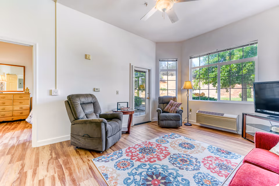 A bright living room with two gray recliner chairs, a red sofa, a colorful patterned rug, a floor lamp, a flat-screen TV on a stand, and large windows showing green trees outside. The room has wood flooring and white walls, with a doorway leading to a bedroom with a wooden dresser and mirror.