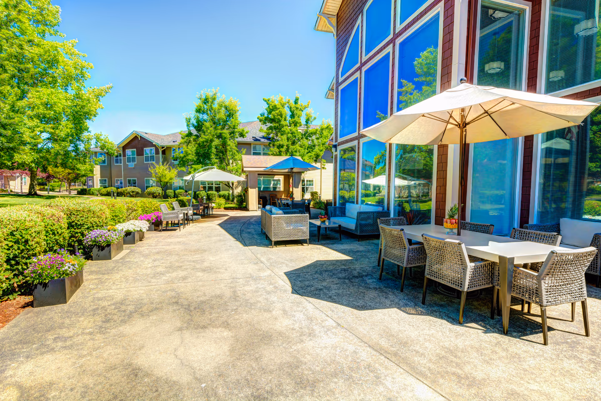 Outdoor patio area at Vineyard Heights Assisted Living with tables, chairs, umbrellas, and lounge seating surrounded by greenery and trees under a clear blue sky.