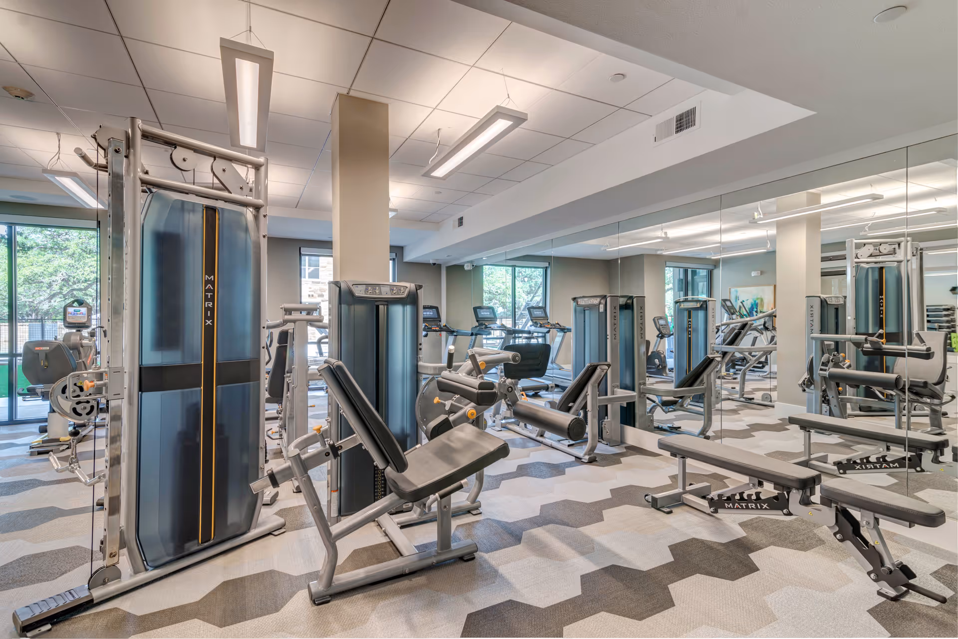 A modern fitness room with various exercise machines including weight machines, benches, and treadmills. The room has large windows letting in natural light and a wall covered with mirrors. The floor has a geometric patterned carpet in shades of gray.