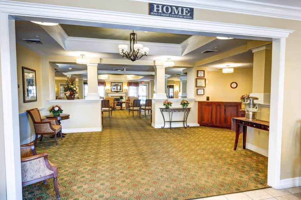 A spacious and well-lit common area in an assisted living facility with patterned carpet, several chairs and tables arranged for seating, floral decorations, and a water dispenser on a wooden table. The walls are painted light yellow, and there is a sign above the entrance that reads 'HOME HOME IS WHERE YOUR STORY BEGINS.'