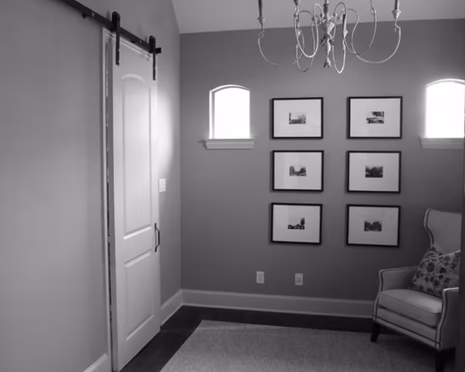 A cozy interior room with a sliding barn door on the left wall, two small arched windows on the back wall, six framed pictures arranged in two columns, and a cushioned armchair with a patterned pillow on the right side. A chandelier hangs from the ceiling.
