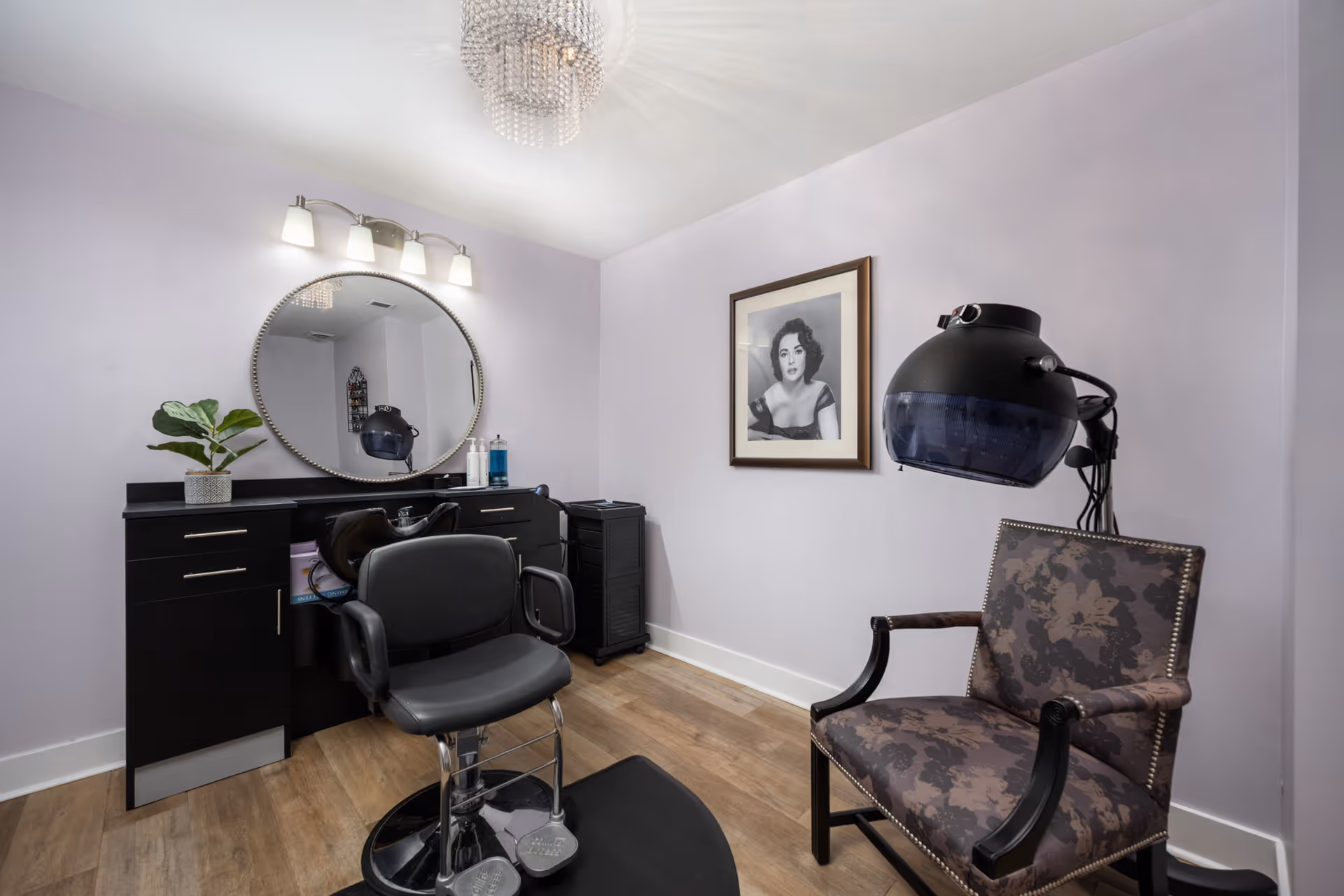 A small salon room with a black salon chair in front of a black counter with a round mirror above it. There is a plant and some hair products on the counter. To the right, there is a vintage hair dryer chair with a hooded hair dryer attached. A framed black and white portrait of a woman hangs on the lavender-colored wall. The floor is wooden, and a chandelier light fixture is visible on the ceiling.