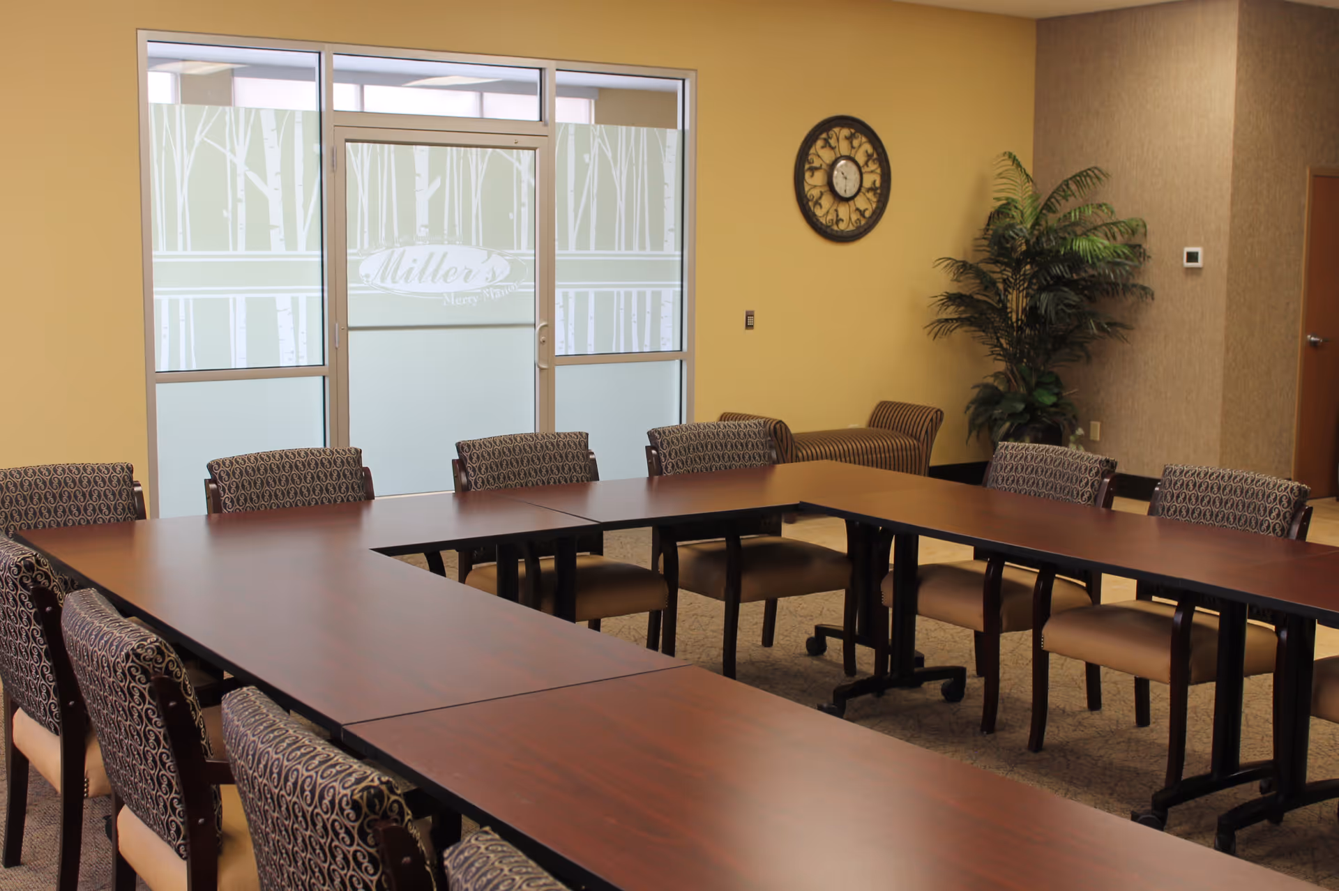 A conference or meeting room with a U-shaped arrangement of wooden tables and patterned chairs. There is a large window with frosted glass featuring a tree design and the text 'Miller's Merry Manor'. A decorative wall clock and a potted plant are visible on the beige walls.