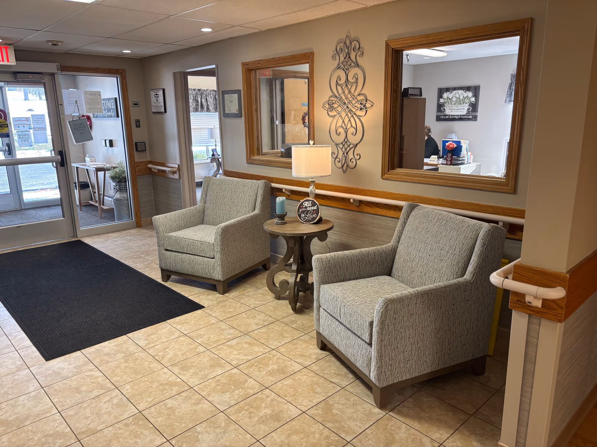 A cozy waiting area inside a healthcare facility with two upholstered armchairs separated by a small round wooden table holding a lamp and a candle. The walls are decorated with two large mirrors and a decorative metal wall art piece. The floor is tiled, and there is a black entrance mat near the glass double doors leading outside. A reception area is visible through the windows on the right side.