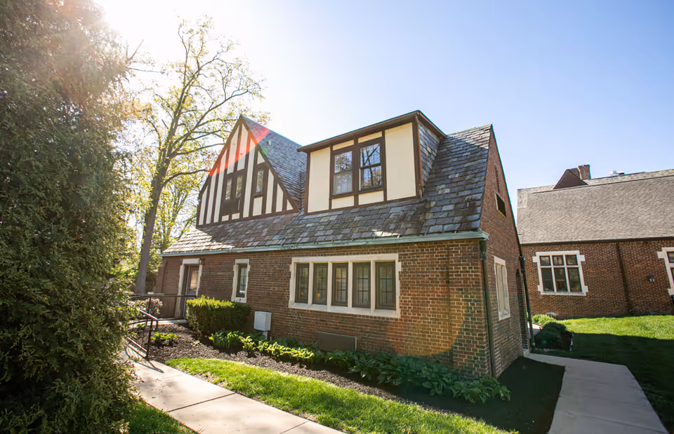 Tudor-style brick building with a steep slate roof, windows, walkway and landscaped lawn under a clear sky.