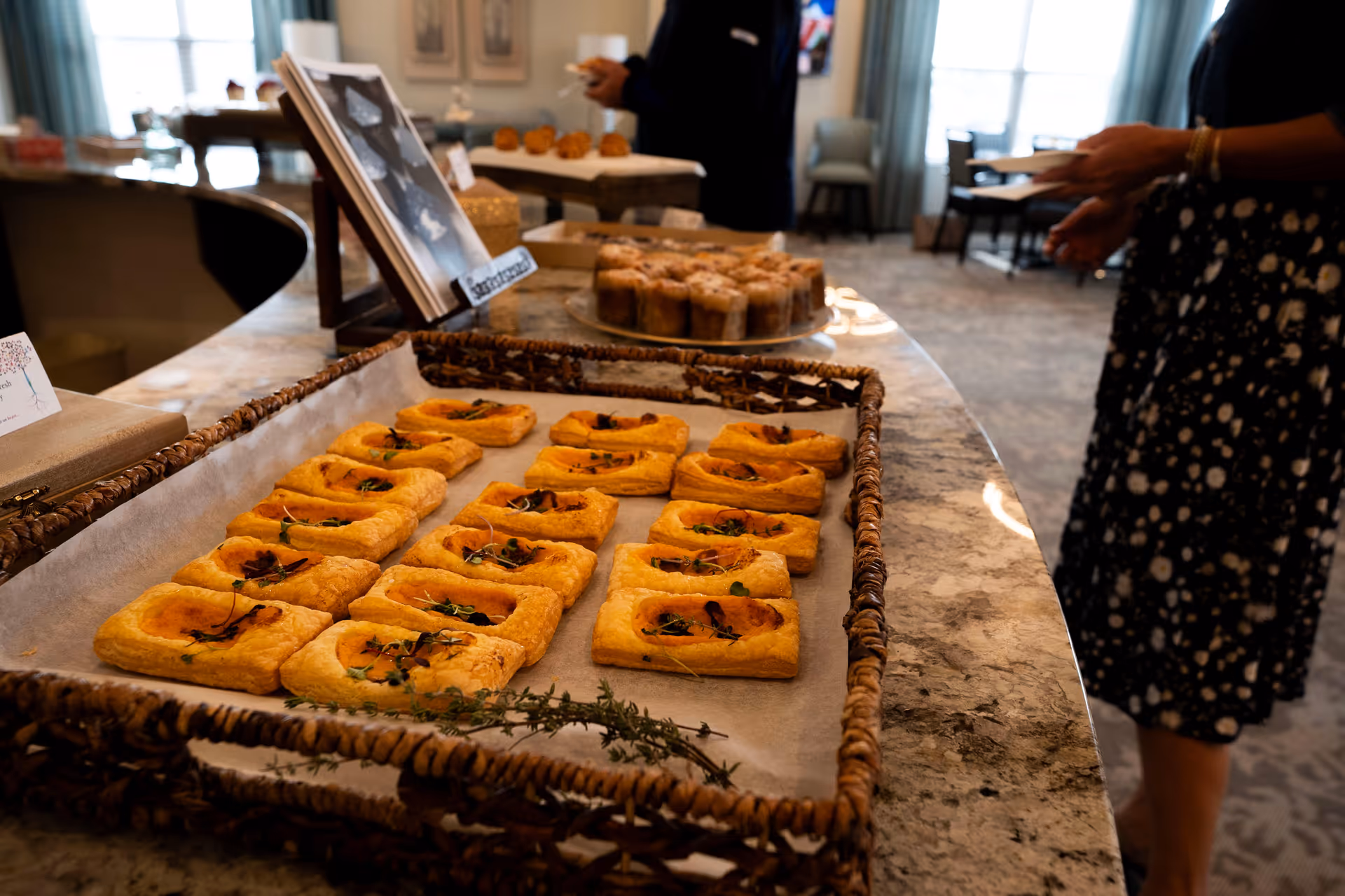 A tray of rectangular pastries garnished with herbs is displayed on a marble countertop in a dining area. In the background, a person is holding a plate, and there are more pastries on a raised stand. The room has large windows with curtains and several chairs and tables.