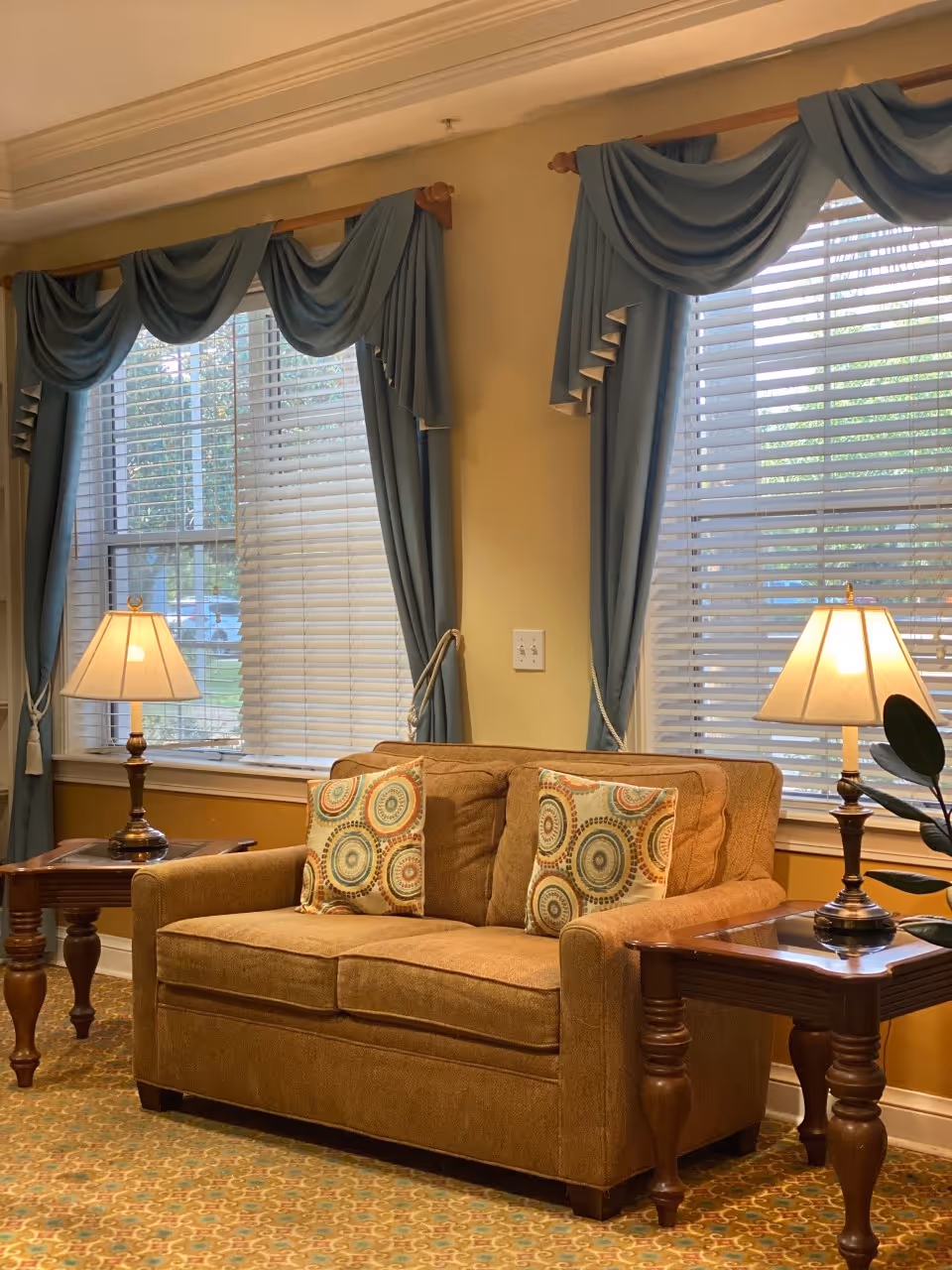 Brown loveseat with patterned pillows between two wooden side tables with lamps in front of windows with blue drapes and blinds.
