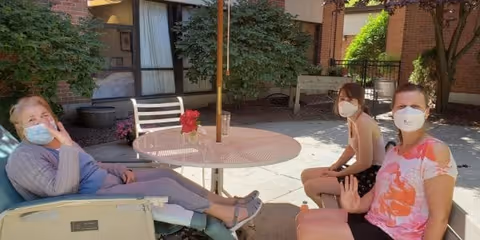 Three people wearing face masks sitting outdoors around a round metal table with a red flower centerpiece. They are in a courtyard area with brick walls, windows, and greenery in the background. Two people are sitting on chairs and one person is reclining on a lounge chair, all waving at the camera.