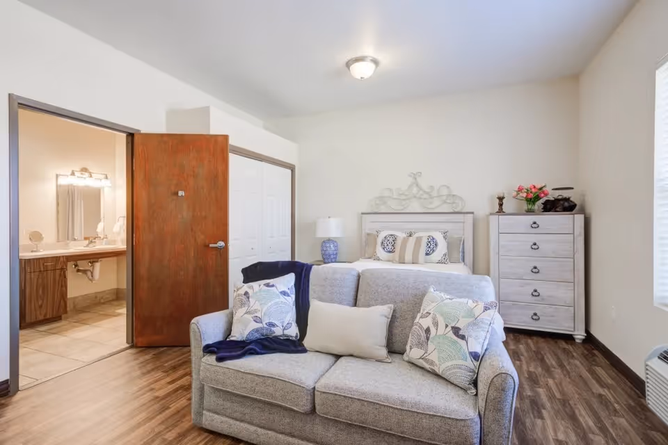 A cozy bedroom with a gray sofa in the foreground adorned with three decorative pillows and a dark blue throw. Behind the sofa is a bed with a decorative metal headboard, pillows, and a bedside lamp on a small table. To the right of the bed is a white chest of drawers with flowers and decorative items on top. The room has wood flooring and light-colored walls. An open door on the left reveals a bathroom with a large mirror and sink.