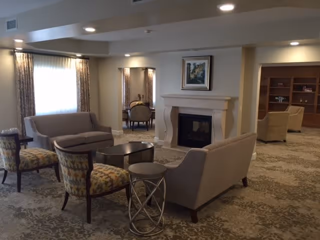 A cozy living room area in an assisted living facility featuring two beige sofas, two patterned armchairs, a small round side table, and a coffee table. The room has a fireplace with a framed painting above it, carpeted floor, and recessed ceiling lights. In the background, there is a dining area with a table and chairs, and a bookshelf with additional seating.