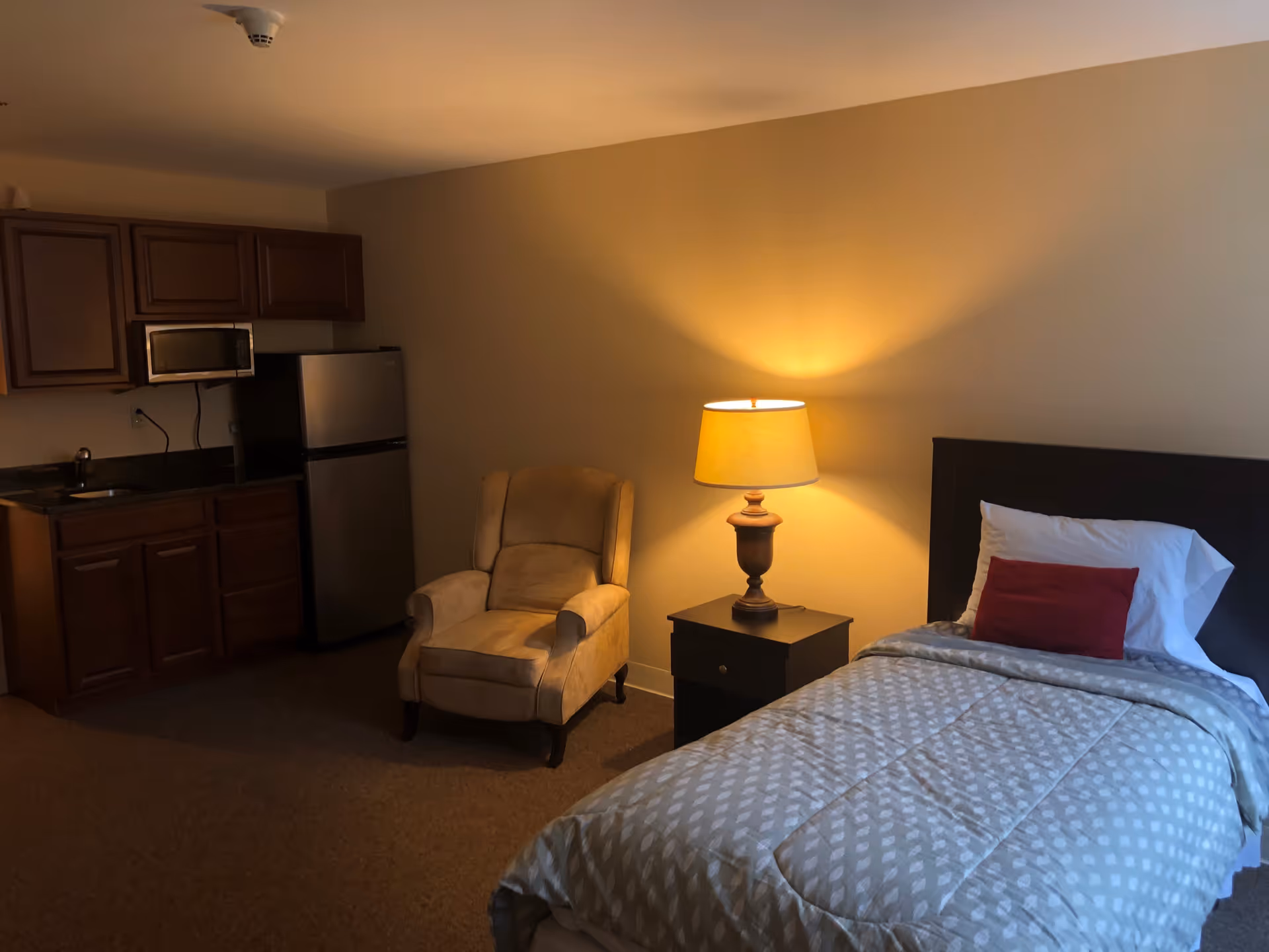 A cozy room featuring a single bed with a patterned gray comforter and a red accent pillow, a wooden nightstand with a lit table lamp, a beige upholstered armchair, and a small kitchenette with wooden cabinets, a microwave, and a stainless steel refrigerator.
