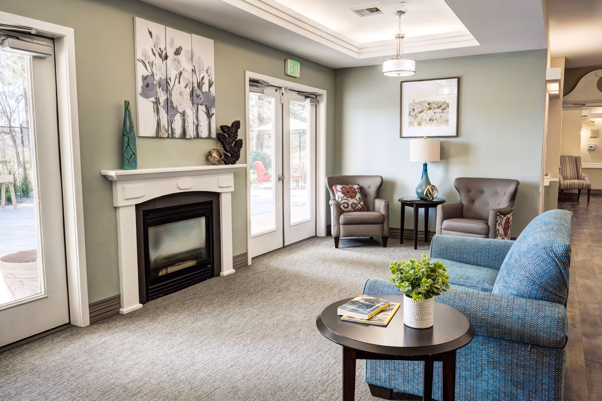 A cozy living room area with a blue upholstered sofa, two gray armchairs with patterned pillows, a round wooden coffee table with a small plant and books, a white fireplace with decorative items on the mantel, and glass double doors leading outside. The walls are painted light green and light blue, with framed artwork and a ceiling light fixture.