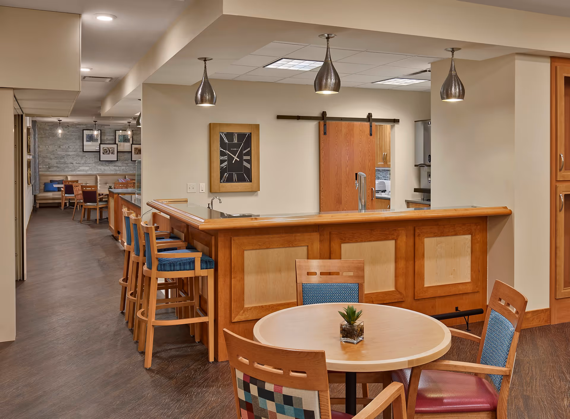 A senior living common area with a wooden bar, high stools, round dining table and chairs, and pendant lights.