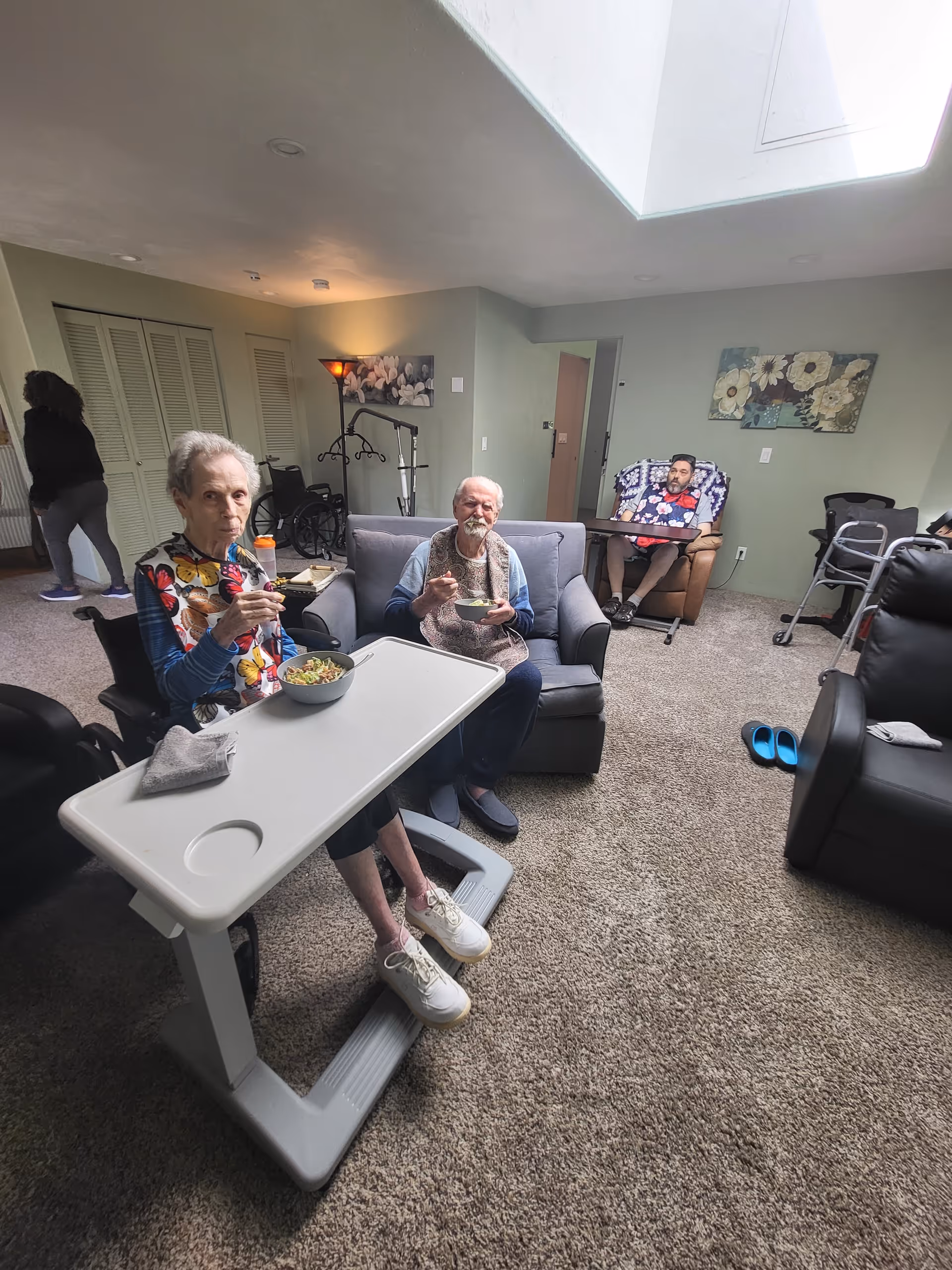 Three elderly individuals sitting in a living room area, two seated on a couch eating from bowls with a tray table in front of one, and another sitting in a recliner with a tray table. The room has carpeted floors, a skylight, floral wall art, and mobility aids like a wheelchair and walker visible.