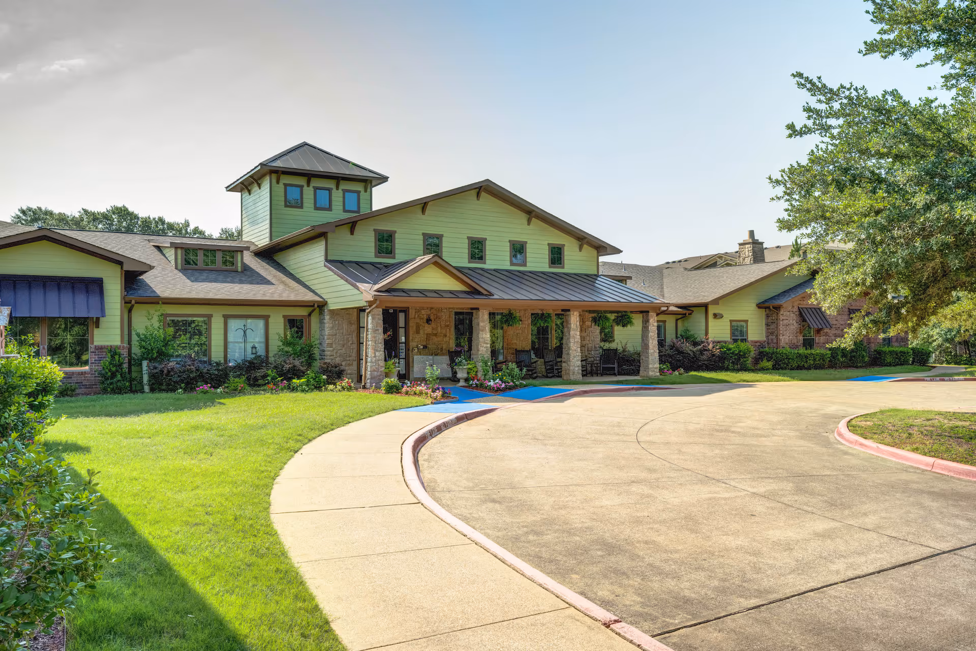 Front exterior of a single-story assisted living building with a covered stone entrance, green siding, landscaped lawn, and a curved driveway.