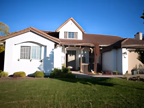 Front exterior view of a single-story house with white walls, brown roof, and a small upper window under a clear blue sky. The house has a porch area with a door and windows, surrounded by green grass and some bushes.