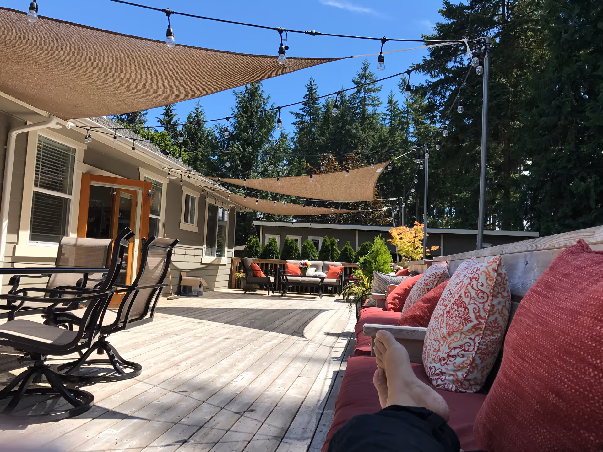 Outdoor patio area with wooden deck, cushioned seating with red and patterned pillows, several chairs around a table, shade sails overhead, string lights hanging above, and trees in the background under a clear blue sky.