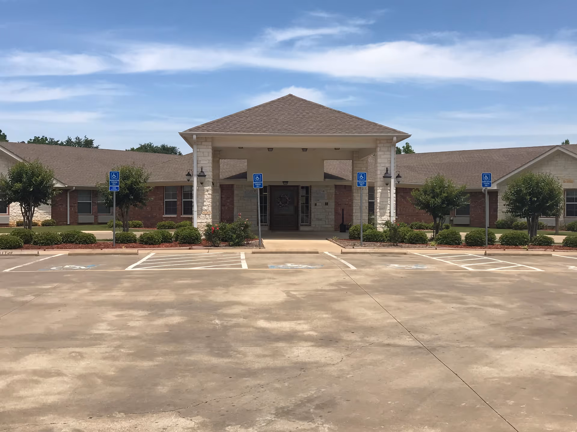 Front exterior view of The Arbors Healthcare and Rehabilitation Center building with a covered entrance, several handicap parking spaces, small trees, and bushes under a partly cloudy sky.
