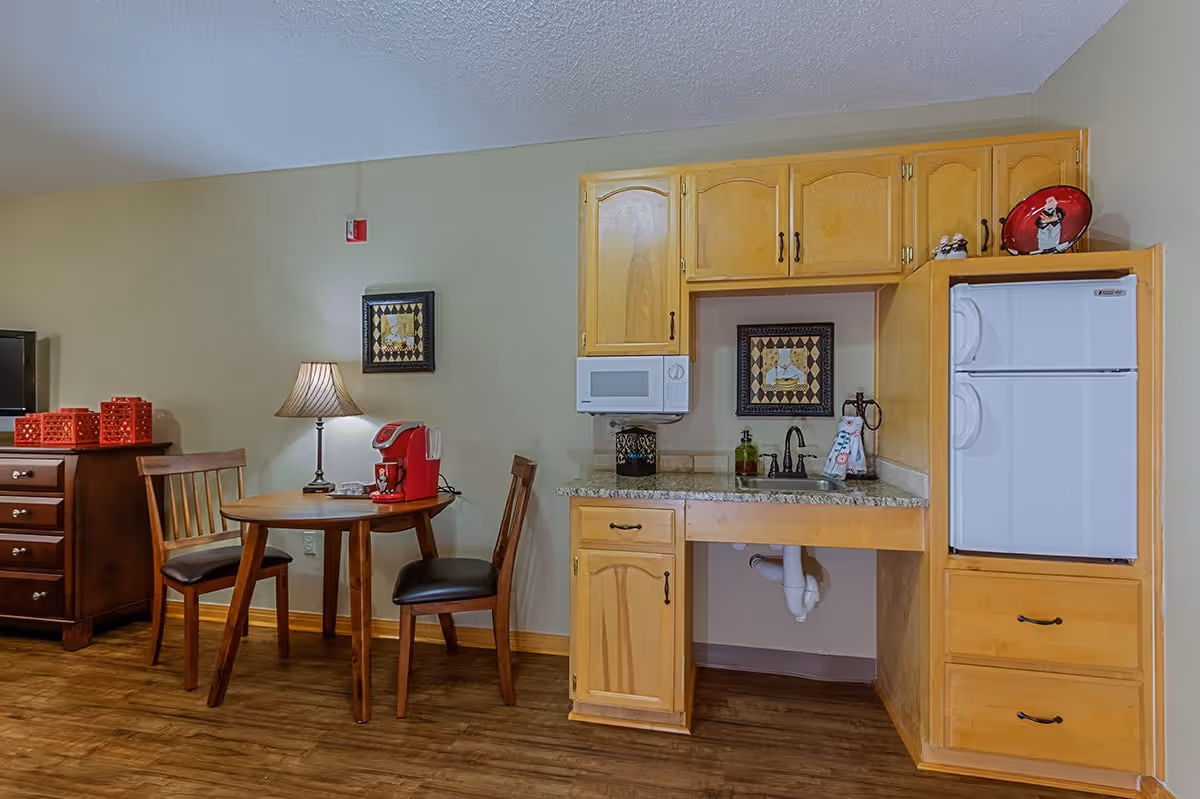 A small kitchenette area with wooden cabinets, a white microwave, a white mini refrigerator, and a granite countertop with a sink. Next to the kitchenette is a round wooden table with two wooden chairs, a table lamp, and a red coffee maker. The floor is wooden, and there are two framed pictures on the wall.