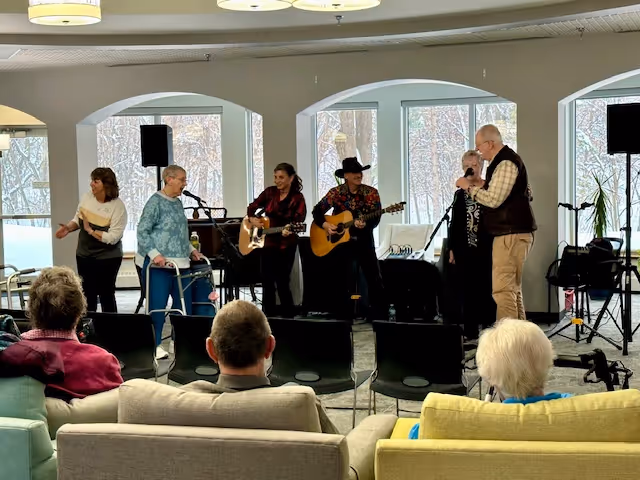 A group of elderly people and musicians in a senior living facility common area. Two musicians are playing guitars and singing, while other seniors are standing or sitting, watching the performance. The room has large windows with a view of trees outside and modern ceiling lights.